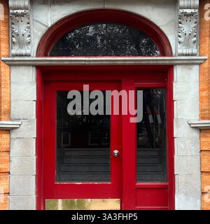 Alte rote Holztür mit Glaspaneelen und einem bogenförmigen Heckfenster, eingerahmt von dekorativen Steindetails an einem historischen Backsteingebäude. Stockfoto