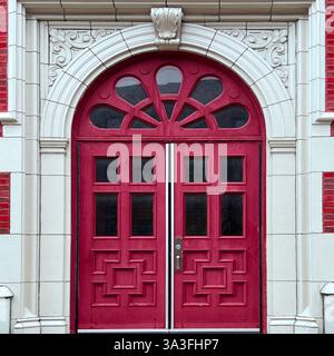 Elegante rote Doppeltüren mit einem Fanlight-Fenster und komplizierten geometrischen Vertäfelungen, eingerahmt von einem dekorativen weißen Steinbogen auf einem historischen roten Backstein. Stockfoto