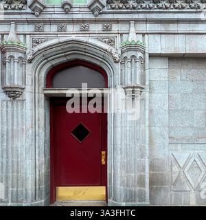 Eine gotisch inspirierte rote Tür mit einem diamantförmigen Fenster, die sich in einem kunstvollen Steinbogen eines historischen Gebäudes befindet. Stockfoto