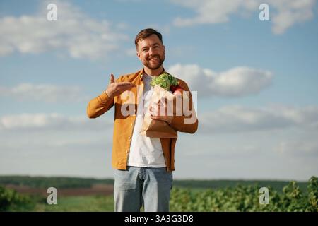 Werfen Sie einen Blick auf die Einkaufstasche mit frischem Kohl und Äpfeln. Ein gutaussehender Mann ist tagsüber auf dem Ackerfeld. Stockfoto