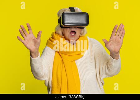 Begeisterte, glückliche Seniorin, die die Headset-Helm-App nutzt, um ein Simulationsspiel zu spielen. 3D 360-Videos in Virtual Reality ansehen. Ältere Großmutter in VR-Brille isoliert auf gelbem Hintergrund. Zukunftstechnologie Stockfoto