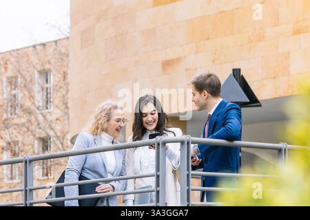 Drei Geschäftsleute führen ein freundliches Gespräch, während sie sich ein Smartphone im Freien ansehen. Sie tragen kluge Geschäftskleidung, smil Stockfoto