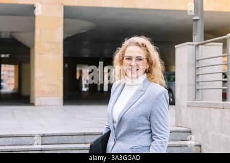 Professionelle Frau mit Brille und grauem Blazer lächelt und hält ein schwarzes Portfolio in einem urbanen Ambiente. Ihre selbstbewusste Einstellung spiegelt Erfolg wider Stockfoto