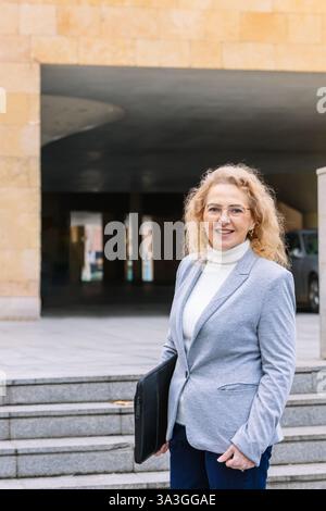 Professionelle Frau, die einen grauen Blazer und eine graue Brille trägt und selbstbewusst lächelt, während sie ein schwarzes Portfolio hält. Sie steht auf einer städtischen Treppe, bereit für Stockfoto