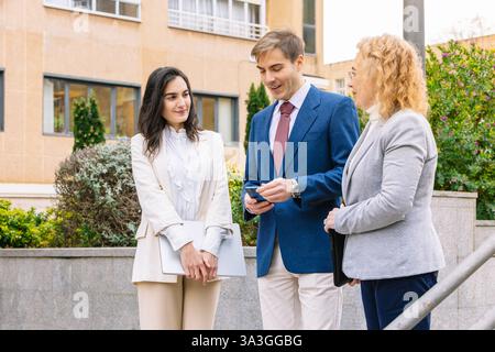 Drei Geschäftsleute führen ein Gespräch in einem urbanen Umfeld. Der Mann in einem blauen Anzug überprüft sein Handy, während die beiden Frauen in Blazern beobachten Stockfoto