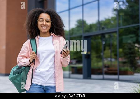 Lächelnde Frau mit lockigem Haar und Brille, die ein Telefon hält, vor einem modernen Gebäude mit einer grünen Seesack. Stockfoto