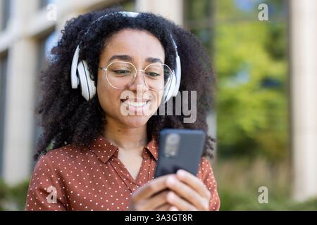 Eine fröhliche junge Frau mit Kopfhörern genießt draußen Inhalte auf ihrem Telefon, trägt ein Polka-Dot-Shirt und eine runde Brille. Stockfoto