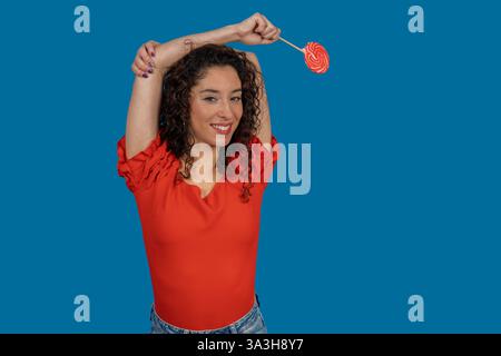 Studio-Porträt einer fröhlichen jungen Frau mit lockigen Haaren, die rote Bonbons auf blauem Hintergrund hält Stockfoto