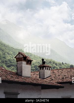Alte Steinschornsteine mit rot gekachelten Terrakottadächern stehen vor einer nebelgrünen Berglandschaft in Venzone, Italien, und schaffen eine zeitlose rustikale Atmosphäre Stockfoto