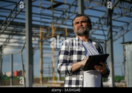 Ernster Gesichtsausdruck. Senior Worker ist in der Nähe des unfertigen Gewächshauses und arbeitet. Stockfoto