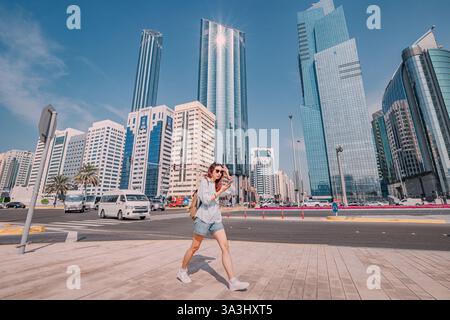16. Januar 2025, Abu Dhabi, VAE: Junge Frau, die die Straße in Abu Dhabi mit Smartphone-App überquert, moderne Wolkenkratzer im Hintergrund Stockfoto
