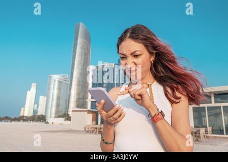 Eine junge Frau, die die Stirn runzelte, während sie Nachrichten auf ihrem Smartphone liest und vor den Wolkenkratzern in der sonnigen Stadt Abu Dhabi steht Stockfoto