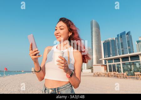 Junge Frau, die am Strand in Abu Dhabi, Vereinigte Arabische Emirate, ihr Handy benutzt und Kaffee trinkt Stockfoto