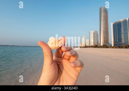 Hand hält eine kleine Muschel am Strand mit der Skyline von Abu Dhabi im Hintergrund, an einem sonnigen Tag Stockfoto