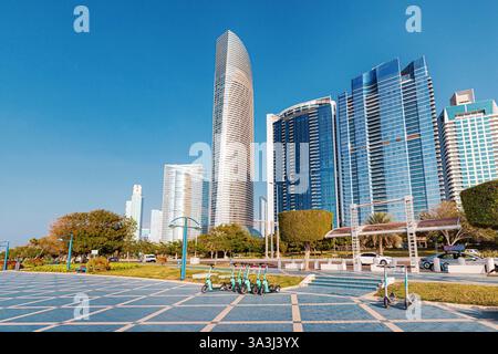 16. Januar 2025, Abu Dhabi, VAE: Elektroroller parken auf einem gefliesten Platz in der Nähe moderner Wolkenkratzer und eines Stadtparks Stockfoto