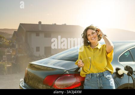 Frau mit gebogenen Haaren, die ein gelbes Hemd trägt, steht neben dem Aufladen des Elektroautos. Windräder im Hintergrund. Stockfoto