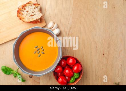 Eine Schüssel Suppe und ein Holzbrett mit Brot. Die Schüssel Suppe steht auf einem Holztisch. Eine Schüssel Tomaten mit Basilikumblättern auf der rechten Seite. Stockfoto
