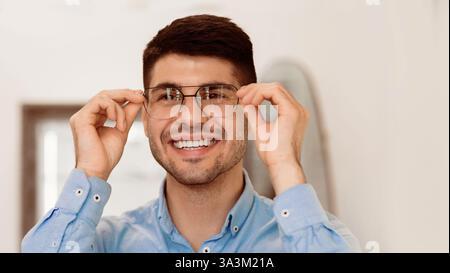 Ein junger Mann passt seine Brille an und lächelt in einem hellen Innenraum Stockfoto