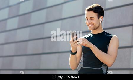 Lächelnder junger Mann in Sportswear mit kabellosen Kopfhörern schaut auf das Smartphone Stockfoto