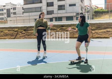 Zwei Freundinnen, eine mit einer Beinprothese, üben Rollschuhlaufen und Skateboarden auf einem farbenfrohen städtischen Sportplatz, zeigen Vielfalt, einschließlich Stockfoto