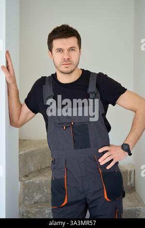 Professioneller Bauarbeiter in schwarzem Overall, der mit überkreuzten Armen in unvollendeter Wohnung steht. Der Mann steht in einem neu gekauften Haus mit Bright Stockfoto
