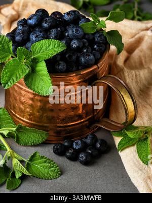 Heidelbeerstillleben. Ein Kupfertopf gefüllt mit frisch gepflückten Heidelbeeren auf einer Fliesenoberfläche mit einem Geschirrtuch und grünen Blättern. Stockfoto