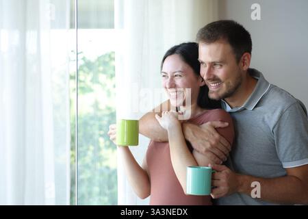 Glückliches, interrassisches Paar, das Kaffee trinkt und zu Hause oder im Hotelzimmer durch das Fenster blickt Stockfoto