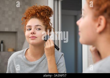 Junge Frau, die Rouge mit dem Pinsel vor dem Spiegel im Badezimmer aufträgt Stockfoto