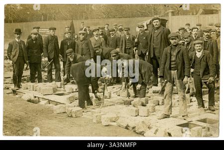 Postkarte aus der Edwardianischen Zeit von Arbeitern, die an der Verlegung von Pflastersteinen arbeiten. Männer aus den 1900er Jahren arbeiten draußen. Sie arbeiten für eine Firma namens The Improved Pavement Company Ltd, siehe Schild, um 1905, 1910 UK Stockfoto