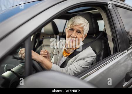 Eine ältere Frau mit kurzen silbernen Haaren lächelt, während sie ihre Brille im Auto hält. Sie trägt ein stilvolles Outfit und zeigt sich während der Fahrt zufrieden Stockfoto