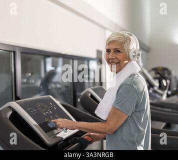 Lächelnde ältere Frau, die im gut ausgestatteten Fitnessstudio auf dem Laufband läuft, Kopfhörer und ein Handtuch um den Hals trägt und sich auf das Training konzentriert. Stockfoto