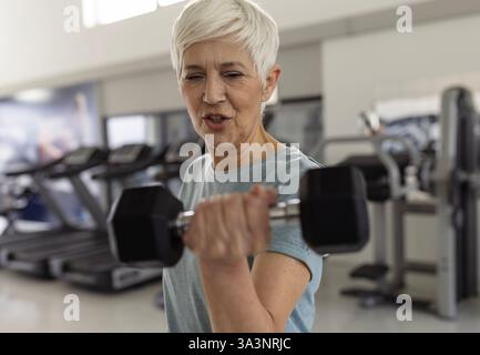 Frau mit kurzen, leichten Haaren hebt im Fitnessstudio eine Hantel und konzentriert sich auf ihr Training. Sie hat es sich zur Aufgabe gemacht, ihre Fitness-Routine in einer hellen Umgebung aufrechtzuerhalten Stockfoto