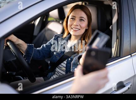Eine Frau in einer Jeansjacke lächelt vom Fahrersitz ihres Autos aus und trifft sich bei Tageslicht mit einer Person, die draußen eine Zahlungskarte hält Stockfoto