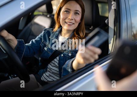Eine Frau in einer Jeansjacke lächelt vom Fahrersitz ihres Autos aus und trifft sich bei Tageslicht mit einer Person, die draußen eine Zahlungskarte hält Stockfoto