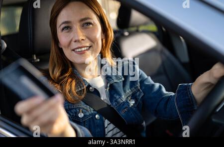 Frau in einer Jeansjacke lächelt vom Fahrersitz ihres Autos auf der rechten Seite, und trifft tagsüber eine Person, die draußen eine Zahlungskarte hält Stockfoto