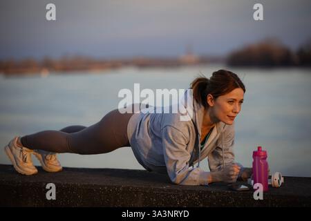 Junge hübsche Frau, die bei Sonnenuntergang an der Wand am Fluss eine Plank-Übung macht. Sie scheint fokussiert und entschlossen zu sein und zeigt ihre Fitnessroutine Stockfoto