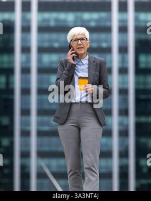 Geschäftsfrau telefoniert, während sie tagsüber außerhalb des modernen Bürogebäudes Kaffee trinkt Stockfoto