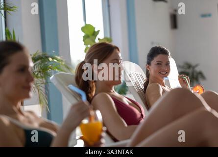 Drei Frauen entspannen sich auf Liegestühlen am Innenpool, schlürfen erfrischende Getränke und genießen die Gesellschaft der anderen am sonnigen Nachmittag Stockfoto