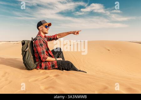 Männlicher Tourist, der nach vorne zeigt, sitzt auf Sanddünen in Wüstenlandschaft mit Rucksack und Sonnenbrille Stockfoto