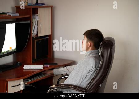 Konzentrierte Jugendliche sitzen in einem bequemen Stuhl am Schreibtisch, tauchen in digitale Studien am Computer ein, umgeben von Büchern und Notizen in einer ordentlichen Innenarbeit Stockfoto