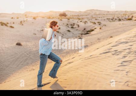 Eine junge Frau lächelt, während sie ein Sandbrett auf ihren Schultern trägt, bei Sonnenuntergang durch die Wüste spaziert und das Abenteuer und die Freiheit annimmt Stockfoto