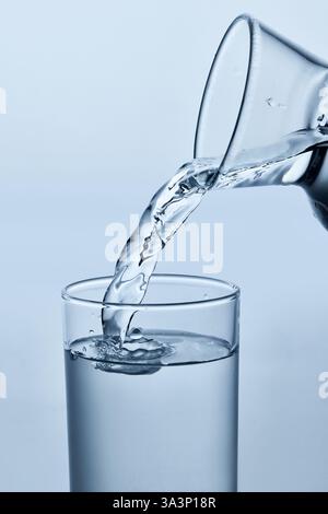 Nahaufnahme des Wassers, das aus dem Krug in klares Glas gegossen wird, mit glattem Strahl und Kräuseln vor hellem Hintergrund. Mock-Up. Stockfoto