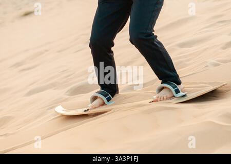 Aus nächster Nähe sehen Sie einen Sandboarder, der eine sonnendurchflutete Düne in der Wüste hinuntergleitet und den Nervenkitzel des Extremsports während des Sonnenuntergangs erfasst Stockfoto