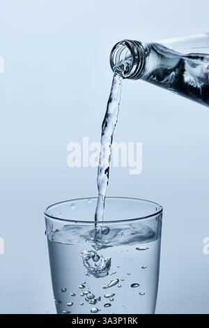 Nahaufnahme des Wassers, das aus der Glasflasche in klares Glas gegossen wird, zeigt fließenden Strom und Spritzer vor hellblauem Hintergrund. Stockfoto