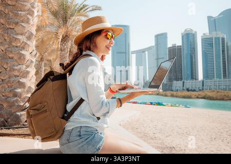 Freiberuflich tätige Frau genießt Online-Arbeit an einem tropischen Strand mit Wolkenkratzern im Hintergrund Stockfoto