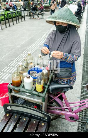 Straßenverkäufer mit konischem Hut und Gesichtsmaske, die Kräutermedizin auf ihrem Fahrradwagen in yogyakarta, indonesien, vorbereitet Stockfoto
