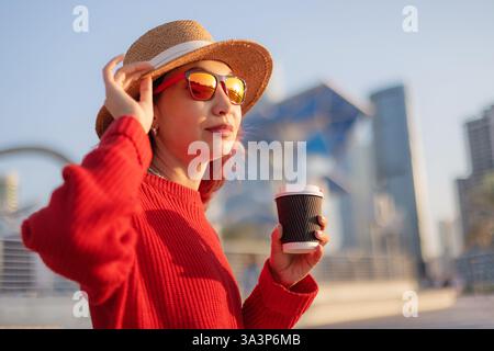 Junge Touristin mit rotem Pullover, Sonnenbrille und Hut, eine Kaffeetasse in der Hand und ihren Hut in Dubai Marina anpassen Stockfoto