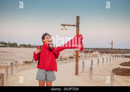 Eine junge Frau in roter Kleidung, die ihren Schal hält, wird bei Sonnenuntergang vom Wind auf einem Fußweg am Strand geblasen Stockfoto