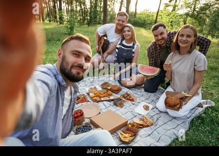 Das Selfie machen, sitzen. Eine Gruppe von Freunden macht ein Picknick auf dem Feld mit Essen in Öko-Boxen. Stockfoto