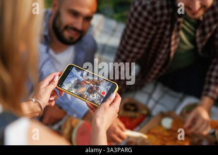 Smartphone in den Händen der Frau, Foto machen. Eine Gruppe von Freunden macht ein Picknick auf dem Feld mit Essen in Öko-Boxen. Stockfoto